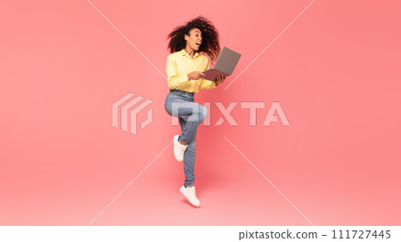 Excited black woman jumps, holding laptop on pink background 111727445