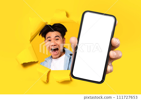 Excited asian guy holding mobile phone breaking through yellow paper Excited asian guy holding mobile phone breaking through yellow paper 111727653