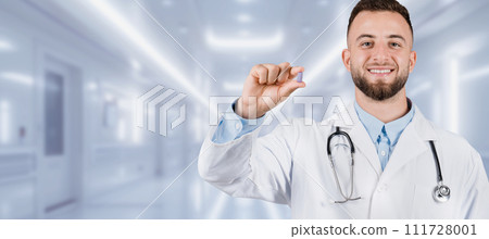 A cheerful young male doctor with a beard is holding a pill between his fingers A cheerful young male doctor with a beard is holding a pill between his fingers 111728001