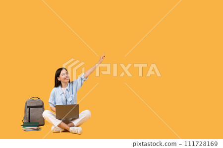 Student lady with laptop pointing upwards at free space sitting beside backpack Student lady with laptop pointing upwards at free space sitting beside backpack 111728169