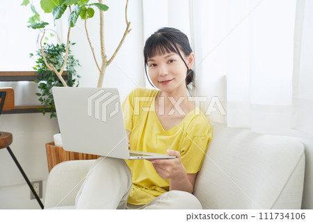 A woman sitting on a sofa by the window and operating a computer 111734106