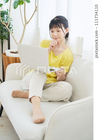 A woman sitting on a sofa by the window and operating a computer A woman sitting on a sofa by the window and operating a computer 111734110