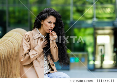 Worried woman coughing outdoors, feeling unwell. Health, sickness, public concern concepts in urban setting. Worried woman coughing outdoors, feeling unwell. Health, sickness, public concern concepts in urban setting. 111735011
