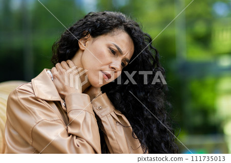 Worried young woman in a stylish beige jacket outdoors, rubbing neck, showing discomfort and pain, with a blurred green background. 111735013