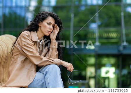 Worried young woman sitting outdoors, holding glasses, deep in thought with a troubled expression, urban backdrop. 111735015