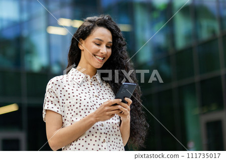 Young, attractive Hispanic businesswoman with curly hair checks her phone with a smile while taking a leisurely walk during her lunch break. 111735017