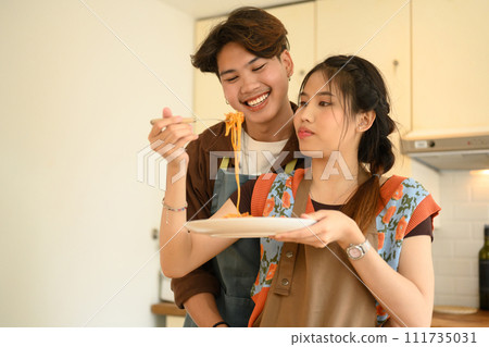 Romantic couple in aprons tasting spaghetti pasta while preparing food together in kitchen 111735031