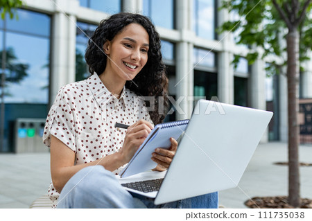 Smiling young Hispanic woman working on her laptop outside an office building, taking notes during a lunch break. 111735038
