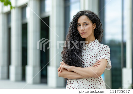 Professional young businesswoman with curly hair standing confidently outside an office, arms crossed during a break. 111735057
