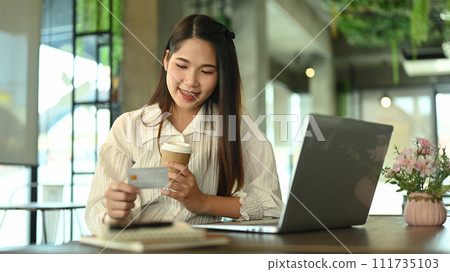 Happy young woman holding paper cup and looking at credit card while shopping online 111735103