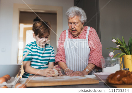 Grandmother with grandson preparing traditional easter meals, kneading dough for easter cross buns. Passing down family recipes, custom and stories. 111735254
