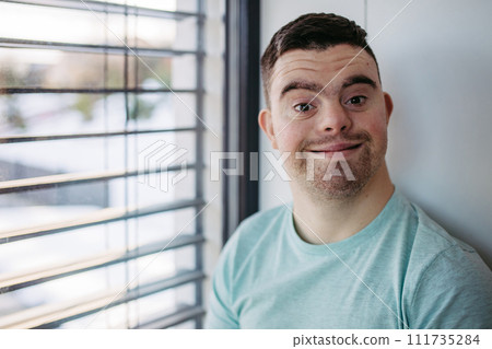Young man with Down syndrome sitting by window, looking at camera. Young man with Down syndrome sitting by window, looking at camera. 111735284