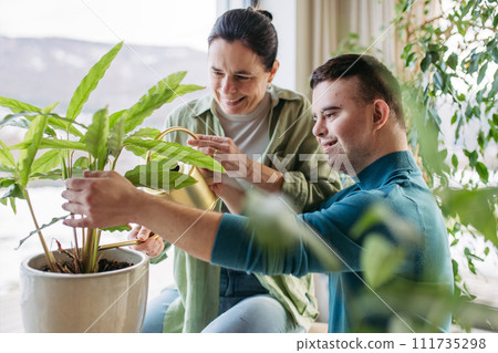 Portrait of young man with Down syndrome with his mother at home, taking care of plants. Concept of love and parenting disabled child. Portrait of young man with Down syndrome with his mother at home, taking care of plants. Concept of love and parenting disabled child. 111735298