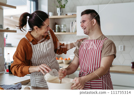 Young man with Down syndrome preparing breakfast with his mother at home, cracking eggs. Morning routine for man with Down syndrome. 111735314