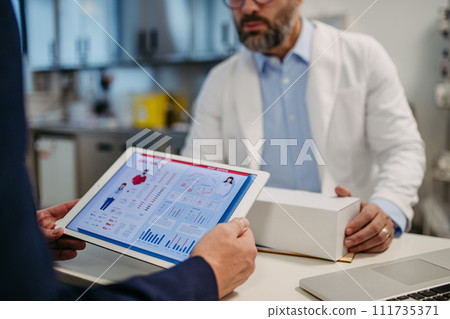 Pharmaceutical sales representative presenting new medication to doctor in medical building, holding tablet with presentation, graphs and statistics. 111735371
