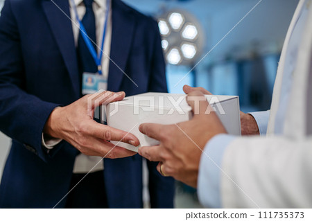 Close up of pharmaceutical sales representative presenting new medication to doctor in medical building, holding box with medication samples, handing it to doctor.. 111735373