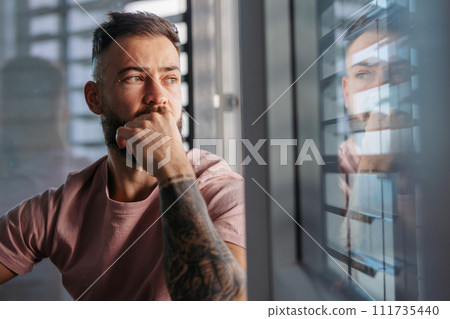 Calm man in pink t-shirt with tattoo on arms looking through window. 111735440