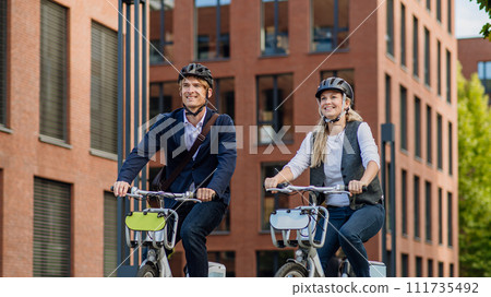 Spouses commuting through the city, talking and walking by bike on street. Middle-aged city commuters traveling from work by bike after a long workday. Spouses commuting through the city, talking and walking by bike on street. Middle-aged city commuters traveling from work by bike after a long workday. 111735492