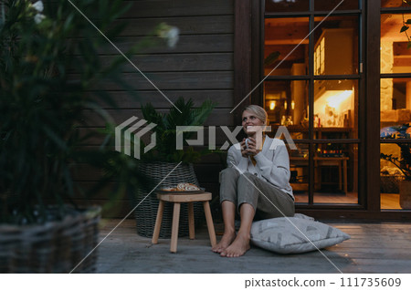 Woman sitting on terrace during dusk, sipping warm tea and enjoying the view. Inside the house, it's warm and inviting, while she is dressed in a cozy sweater, sitting on soft pillows. Hygge 111735609