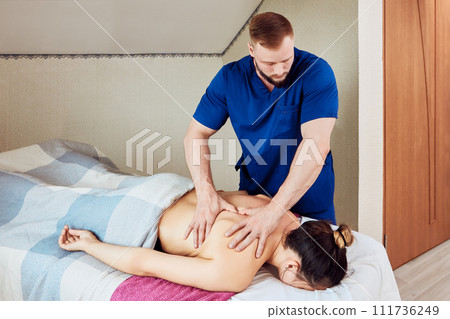 White bearded man in blue robe massages back of European woman lying face down on massage table. White bearded man in blue robe massages back of European woman lying face down on massage table. 111736249