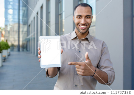 Portrait of a smiling young hispanic man standing near an office building, holding a mock-up tablet and pointing at a white screen. Portrait of a smiling young hispanic man standing near an office building, holding a mock-up tablet and pointing at a white screen. 111737388