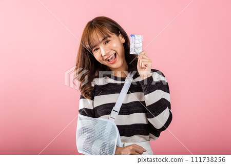Smiling Asian woman with a broken arm wears an arm splint and handles emergency medical bills using a credit card. Happy female with a sling-supported hand in a studio shot on pink background. 111738256