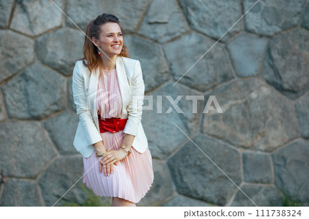 smiling modern woman in pink dress and white jacket in city 111738324