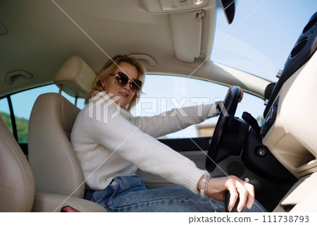 A blonde woman in a white sweater and jeans is driving. Happy woman sitting in a car with a white interior. 111738793