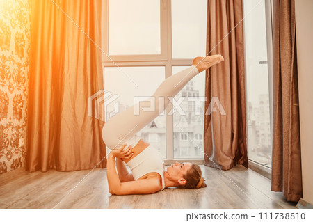 Young athletic attractive woman practicing yoga. Works out at home or in a yoga studio, sportswear, white pants and a full-length top indoors. Healthy lifestyle concept Young athletic attractive woman practicing yoga. Works out at home or in a yoga studio, sportswear, white pants and a full-length top indoors. Healthy lifestyle concept 111738810