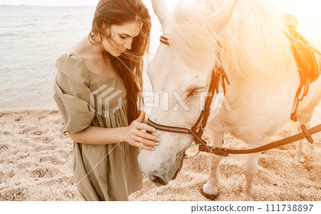 A white horse and a woman in a dress stand on a beach, with the 111738897