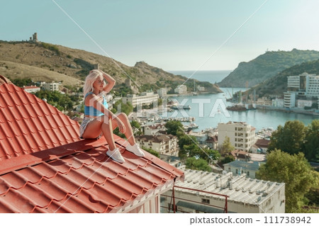 Woman sits on rooftop, enjoys town view and sea mountains. Peaceful rooftop relaxation. Below her, there is a town with several boats visible in the water. Rooftop vantage point. 111738942