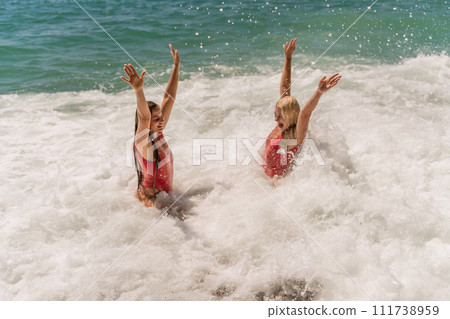 Women ocean play. Seaside, beach daytime, enjoying beach fun. Two women in red swimsuits enjoying themselves in the ocean waves and raising their hands up. Women ocean play. Seaside, beach daytime, enjoying beach fun. Two women in red swimsuits enjoying themselves in the ocean waves and raising their hands up. 111738959