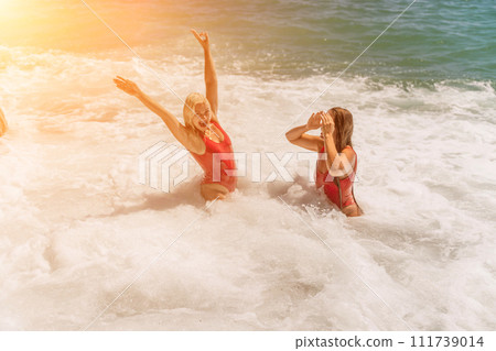 Women ocean play. Seaside, beach daytime, enjoying beach fun. Two women in red swimsuits enjoying themselves in the ocean waves and raising their hands up. 111739014