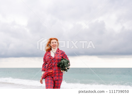 Sea Lady in plaid shirt with a christmas tree in her hands enjoys beach. Coastal area. Christmas, New Year holidays concep 111739050