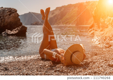 woman sea travel Tanned middle-aged woman with long hair and a white bathing suit. He sits on the seashore in a large sun hat with his back and looks at the sea. 111740268