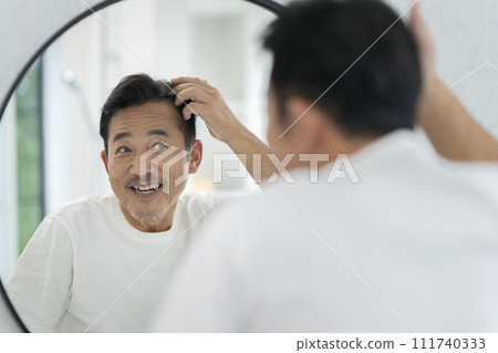 Smiling man in his 50s touching his hair while looking in the mirror Smiling man in his 50s touching his hair while looking in the mirror 111740333