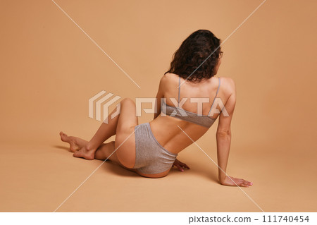 Rear view photo of young brunette woman sitting on floor and posing in lingerie against sandy color studio background. Concept of beauty and fashion. Rear view photo of young brunette woman sitting on floor and posing in lingerie against sandy color studio background. Concept of beauty and fashion. 111740454