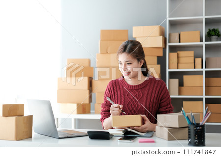 Startup SME small business entrepreneur of freelance Asian woman wearing apron using laptop and box to receive and review orders online to prepare to pack sell to customers, online sme business ideas. 111741847