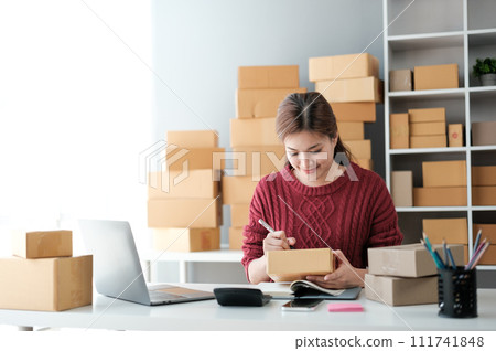 Startup SME small business entrepreneur of freelance Asian woman wearing apron using laptop and box to receive and review orders online to prepare to pack sell to customers, online sme business ideas. 111741848