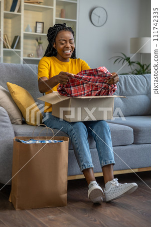 Smiling and happy woman looking contented while opening a box with clothes Smiling and happy woman looking contented while opening a box with clothes 111742335