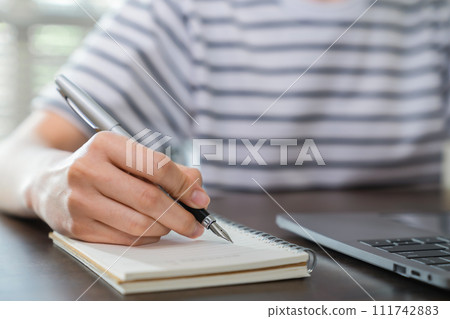Close-up of hand writing on blank notebook on table. Close-up of hand writing on blank notebook on table. 111742883