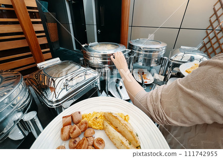 A lady at the hotel buffet is selecting food from a warming machine to place on her plate. A person arranging food on a plate from their own perspective. A lady at the hotel buffet is selecting food from a warming machine to place on her plate. A person arranging food on a plate from their own perspective. 111742955