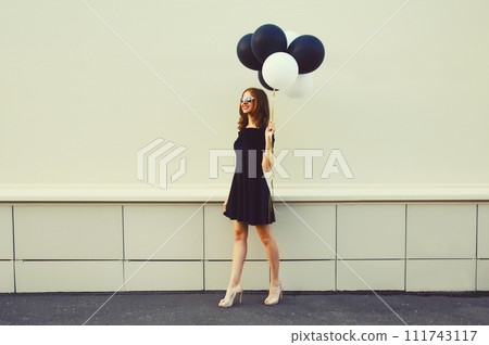 Beautiful happy young woman with bunch of black and white balloons in summer dress on city street 111743117