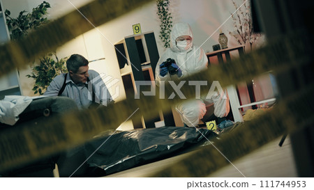View through the yellow police tape at the crime scene, with a forensic expert and a detective collecting evidence View through the yellow police tape at the crime scene, with a forensic expert and a detective collecting evidence 111744953