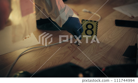 A forensic expert in protective gear is collecting evidence into a plastic bag after taking photos 111744994