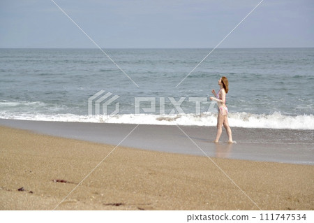 A young woman wearing a bikini swimsuit walking along the beach 111747534