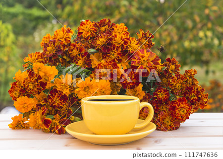 Summer leisure activities concept; yellow cup of coffee and bunch of orange marigold flowers on a wooden table in green garden 111747636