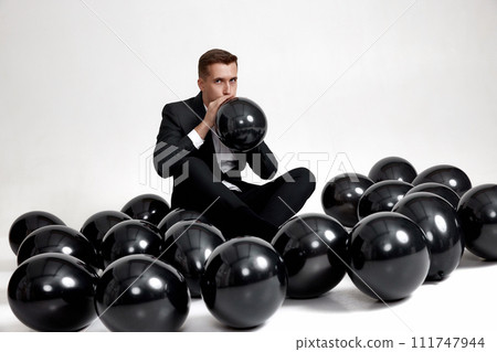 handsome man in black jacket blowing black air balloon on white studio background. new year party handsome man in black jacket blowing black air balloon on white studio background. new year party 111747944