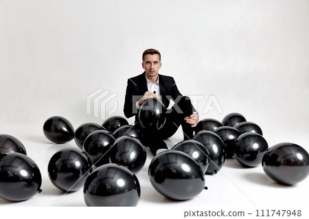 elegant caucasian man in suit tuxedo sitting on studio floor with a lot of black air balloons 111747948