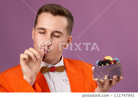 handsome man in orange jacket eating tasty cake on purple background. celebrating birthday, enjoying holidays tasty food 111747991